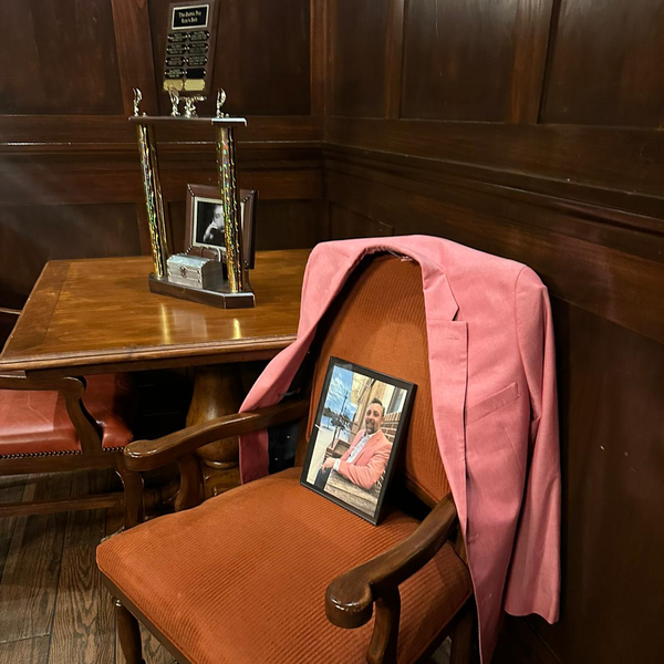 Chair with a pink jacket draped over it, next to a wooden table with a trophy and photo frame in a room with wooden walls.