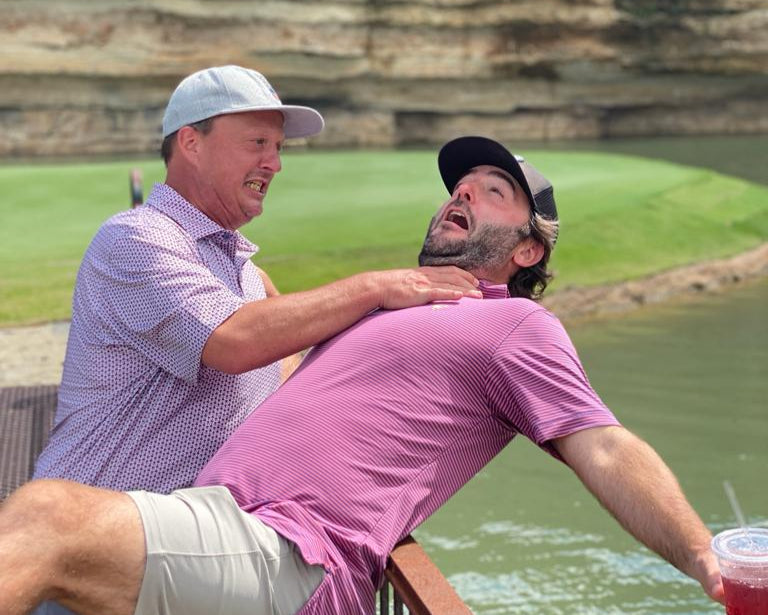 Two men by a body of water with one holding a drink.